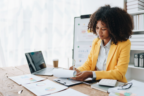 Une femme en blazer jaune examine des documents à un bureau avec un ordinateur portable dans un bureau lumineux