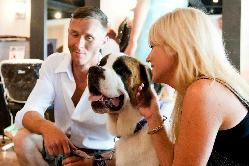Man and woman petting a large St. Bernard dog indoors; man smiles, woman strokes dog's head.