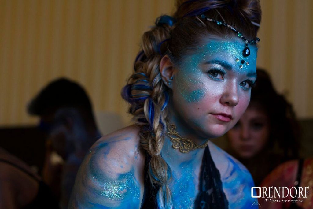Woman with blue body paint and braided hair, wearing a necklace and headpiece.
