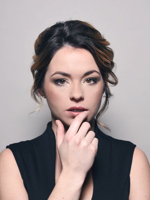 Woman with brown hair, finger to lips, looking at the viewer. Wearing a black sleeveless top.