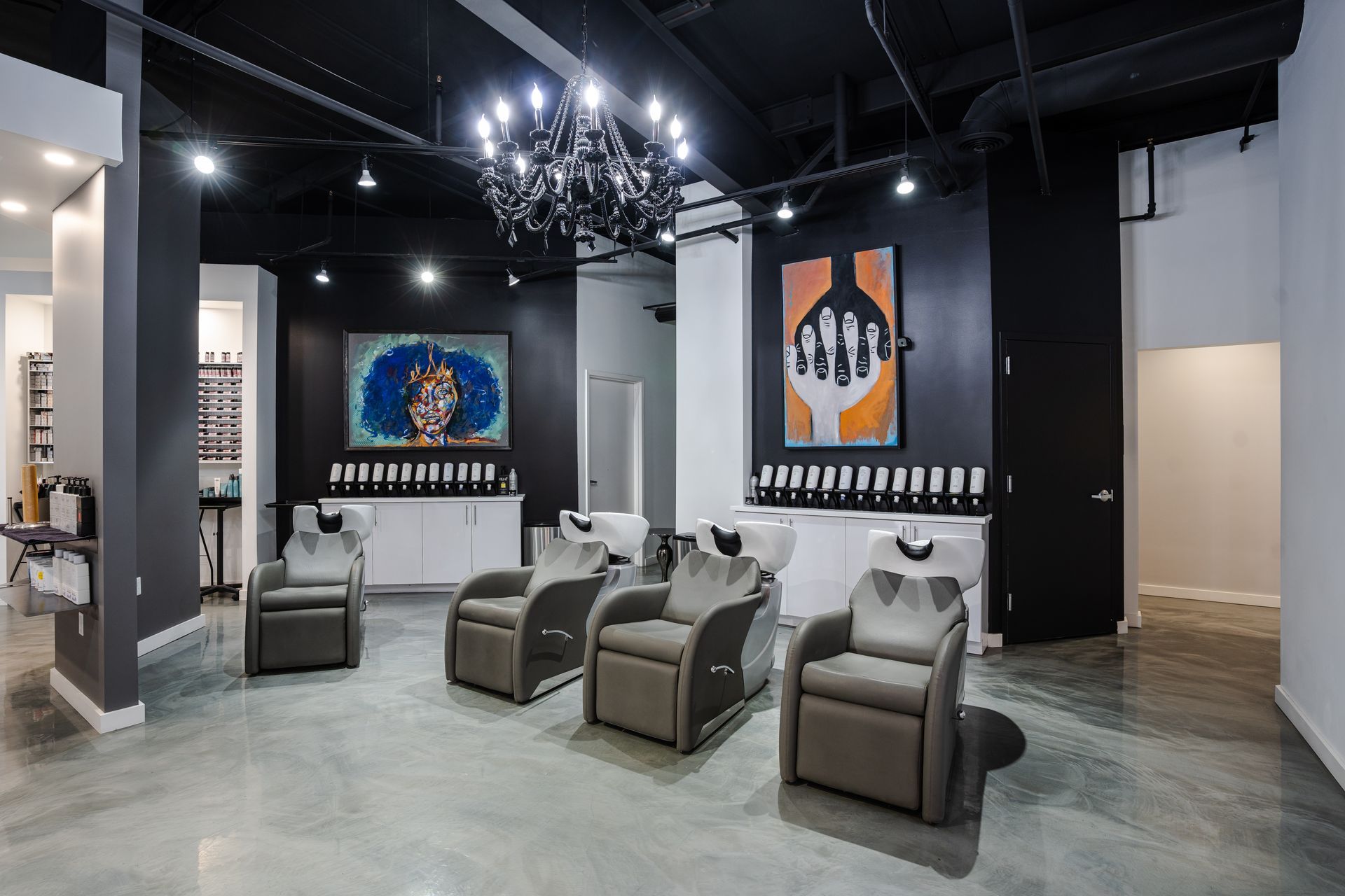 Hair salon interior with dark walls, gray chairs, artwork, and a chandelier.