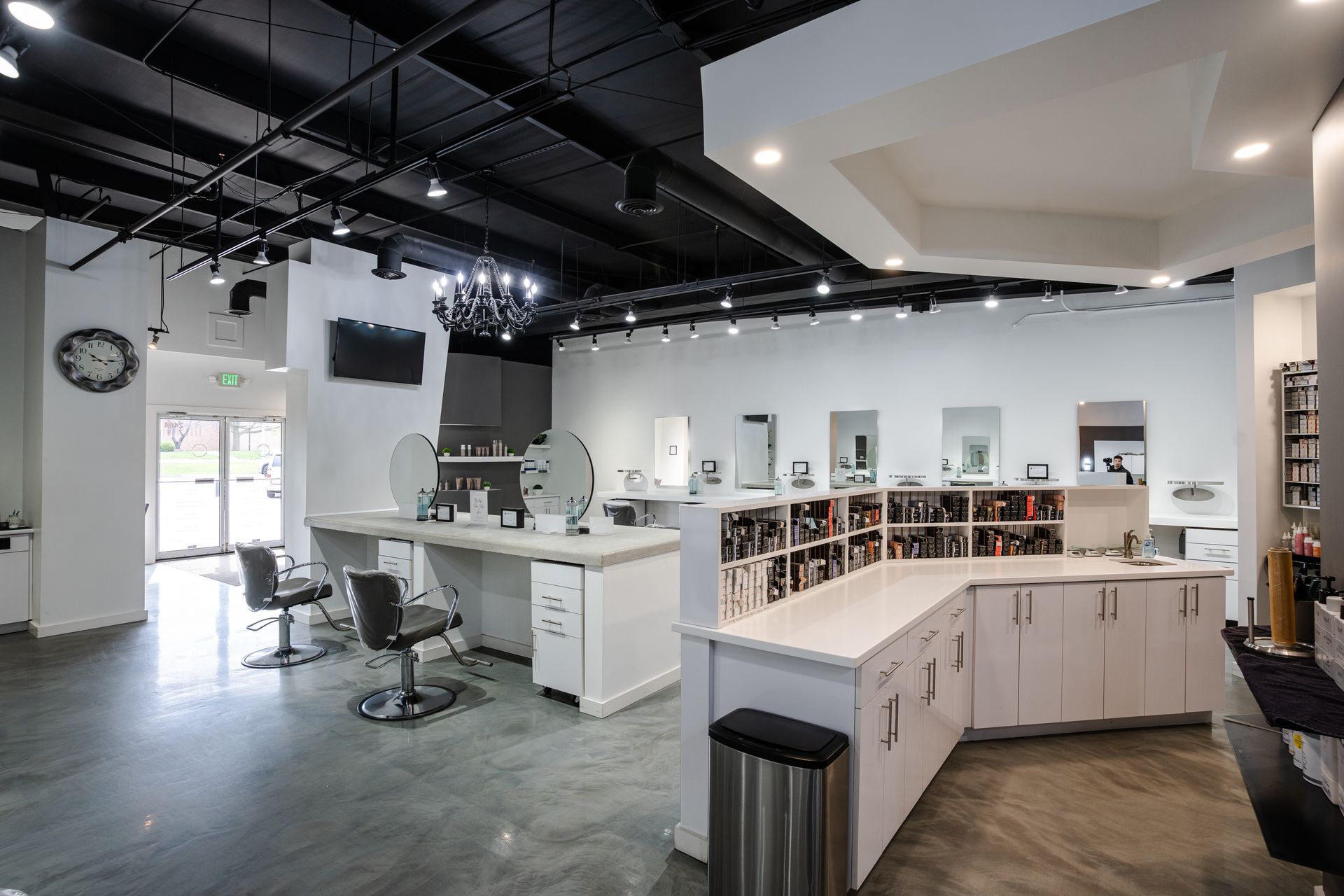 Hair salon interior with white counters, mirrors, and black chairs. Gray floors, black ceiling.