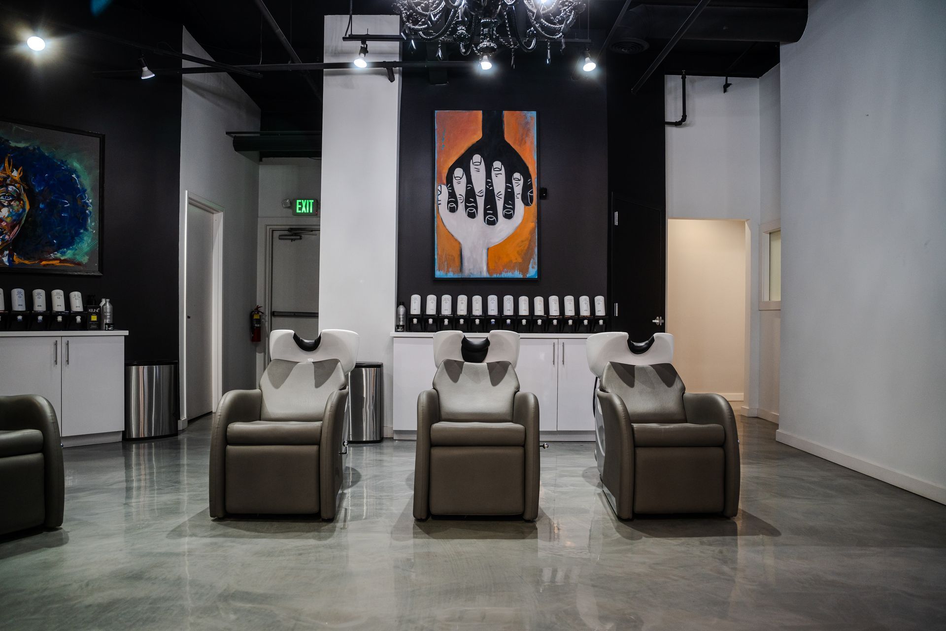 Hair salon interior with shampoo stations. Grey chairs, glossy floor, black accent walls, and artwork.