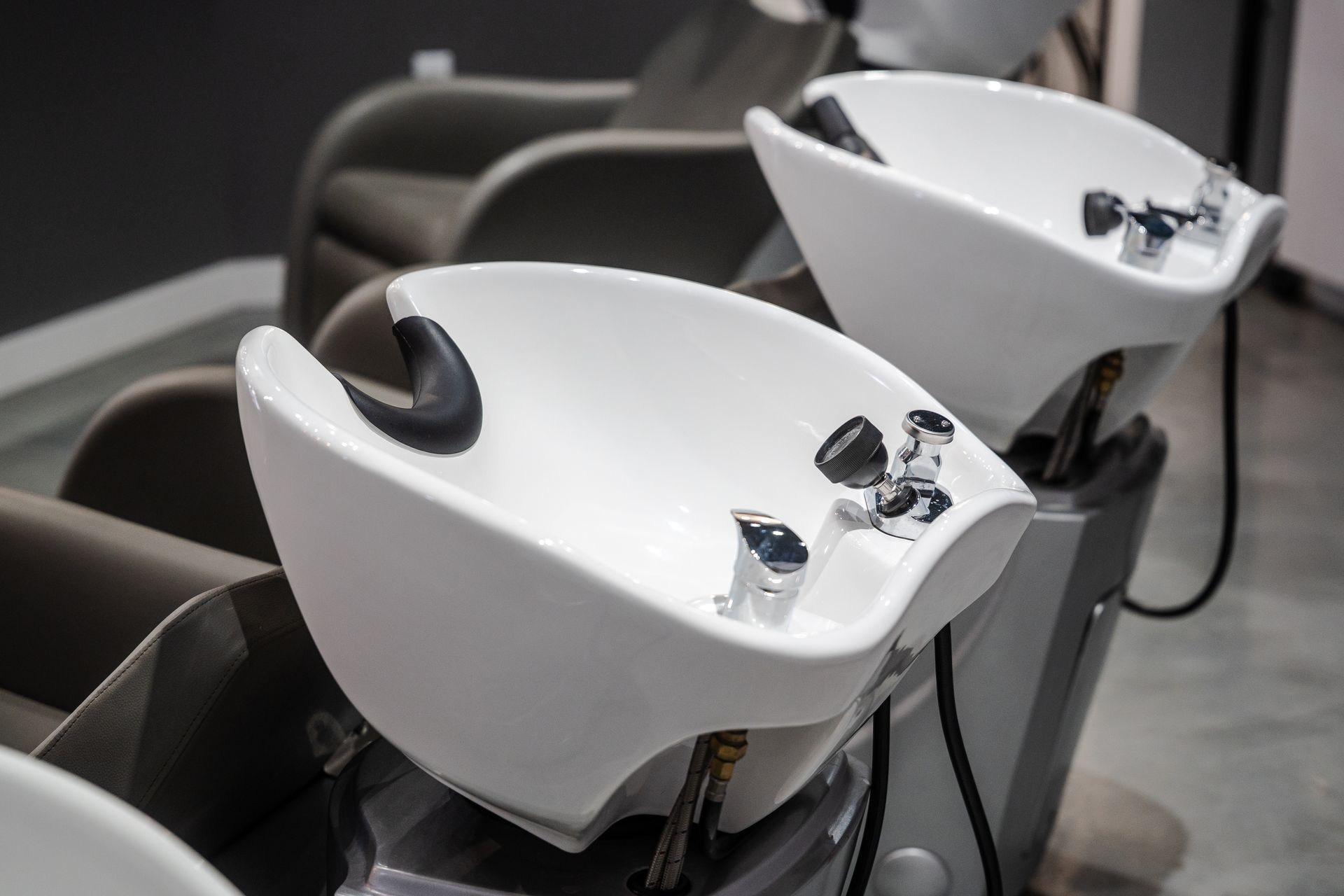 Two white salon shampoo bowls with black neck rests; gray chairs in background.