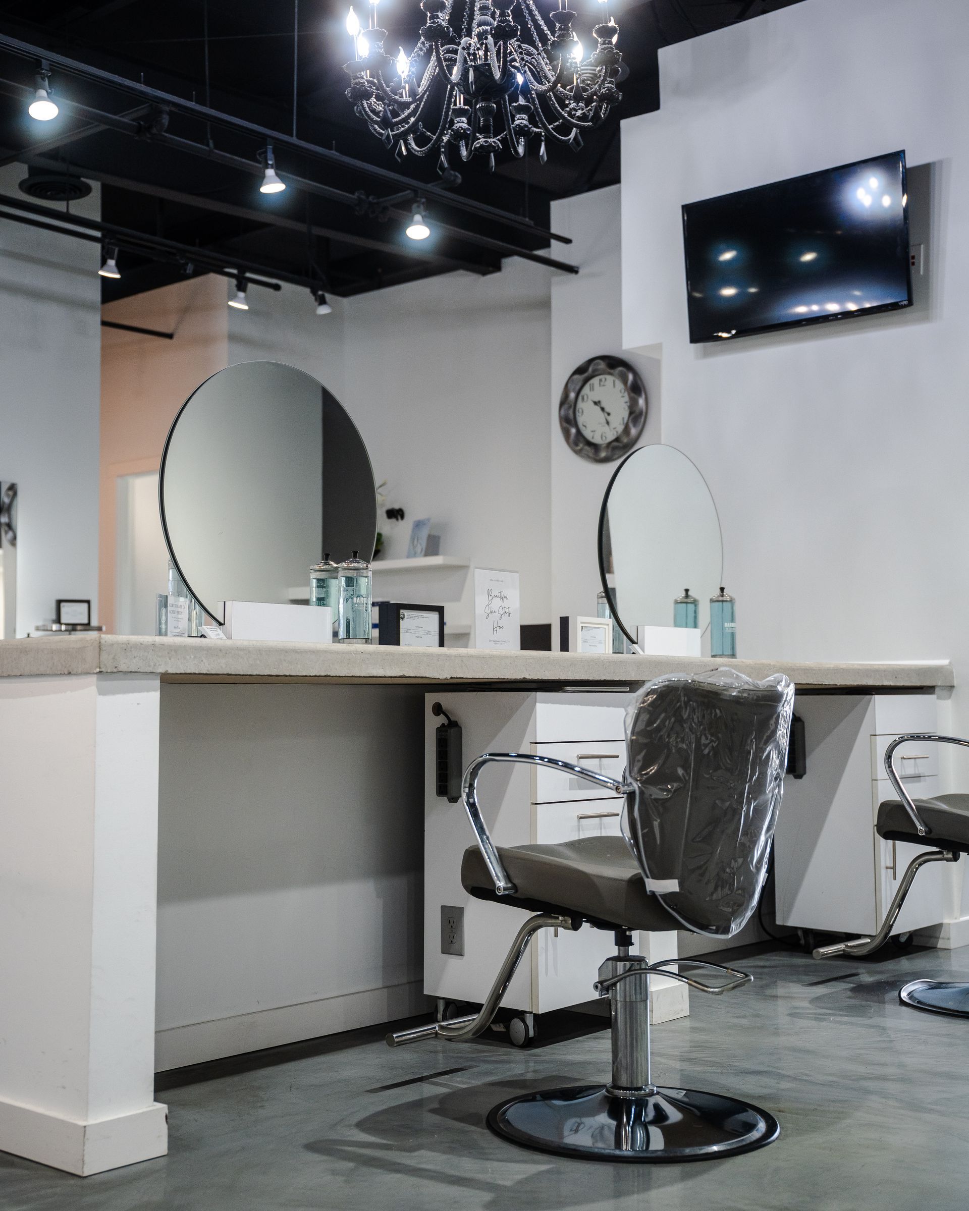Hair salon interior with styling stations, large mirrors, a television, and a chandelier.
