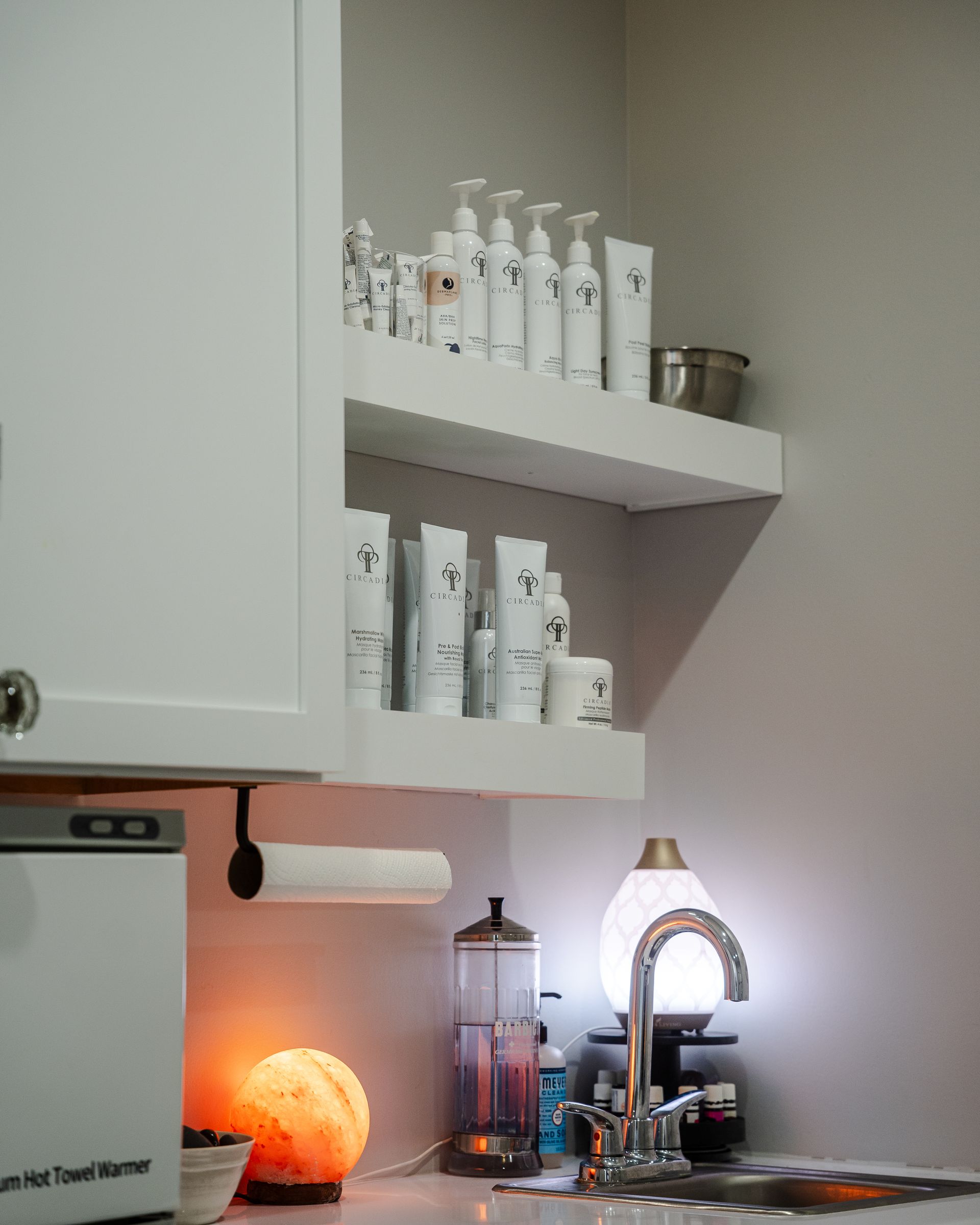 White shelves with skincare products above a sink, illuminated by a lamp and a salt rock.