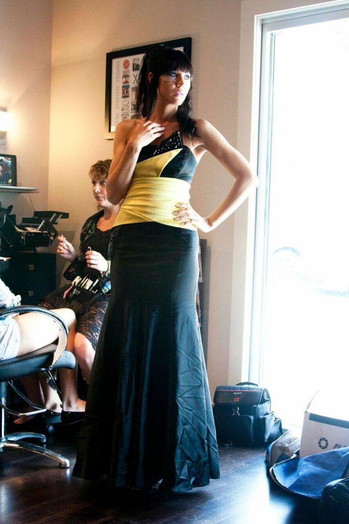 Woman in a yellow and black gown poses indoors with another person in the background.