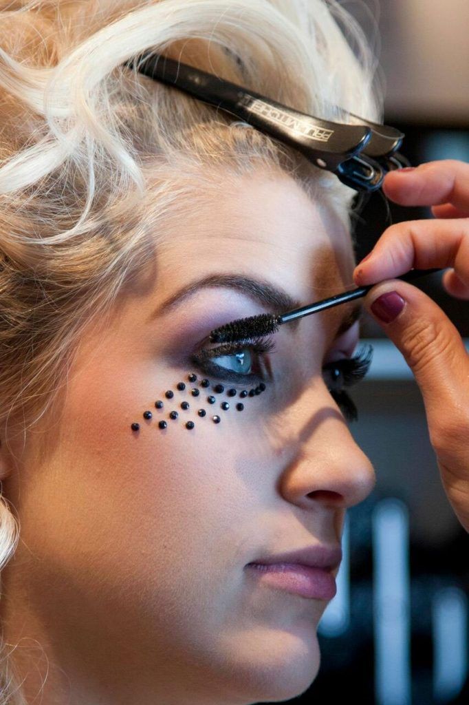 Woman having mascara applied, side profile; elaborate eye makeup with black embellishments.
