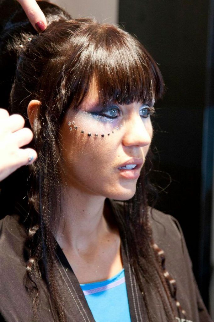 Woman with dark hair in a hairstyle with braids and artistic makeup, being styled with someone's hand touching the hair.