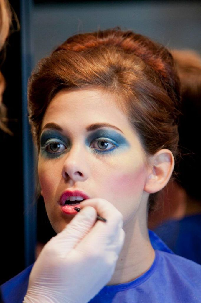 Woman with red hair, blue eyeshadow, and pink blush. Someone applies lipstick to her mouth.