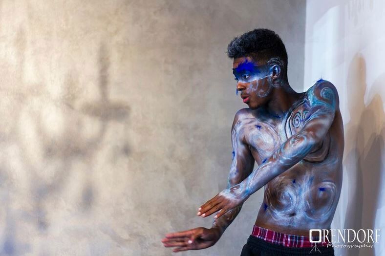 Person with blue and white body paint, arms outstretched, standing in front of a textured wall.
