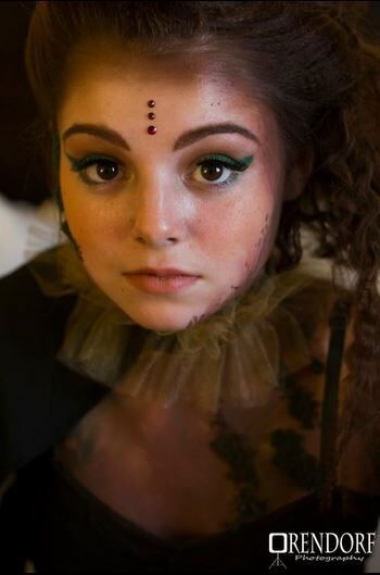 Woman with artistic makeup, three forehead gems, ruffled collar, looking forward.