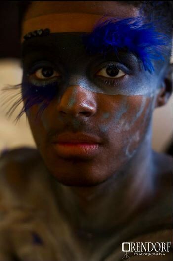 Man with dark face paint, blue feather accents, gazing directly at camera.