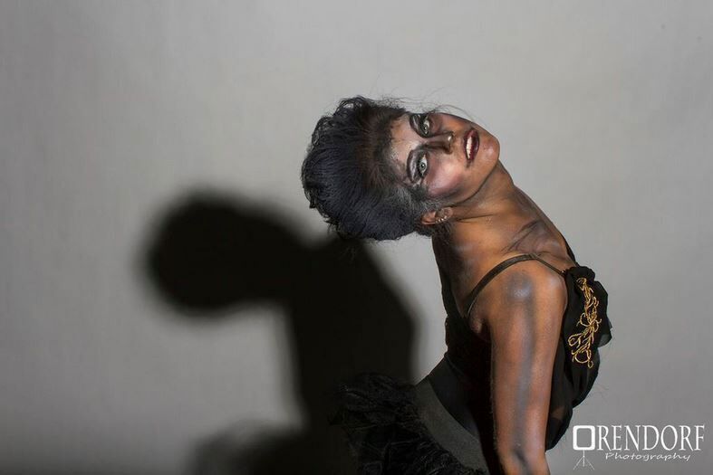 Woman in black costume, arched back, looking up with dramatic makeup and lighting.