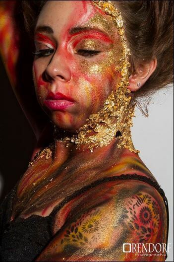 Woman with fire-themed face and body paint, including gold leaf. Eyes closed, looking down. Dark background.
