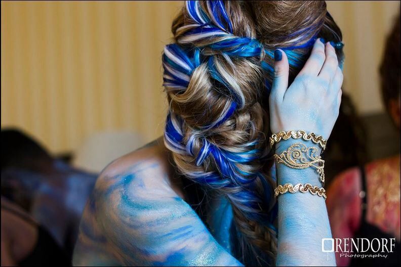 Woman with blue body paint and braided hair with blue streaks, wearing gold bracelets.