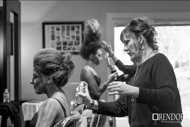 Two stylists working on clients in a salon, one spraying hair and another styling an updo.