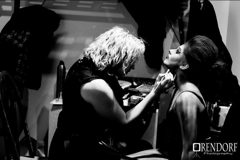 Makeup artist applies makeup to a person’s face, likely a bride, in a room with clothes hanging in the background.