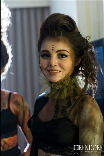 Woman with dark makeup, wearing a green collar and a black outfit, smiling, in a room.