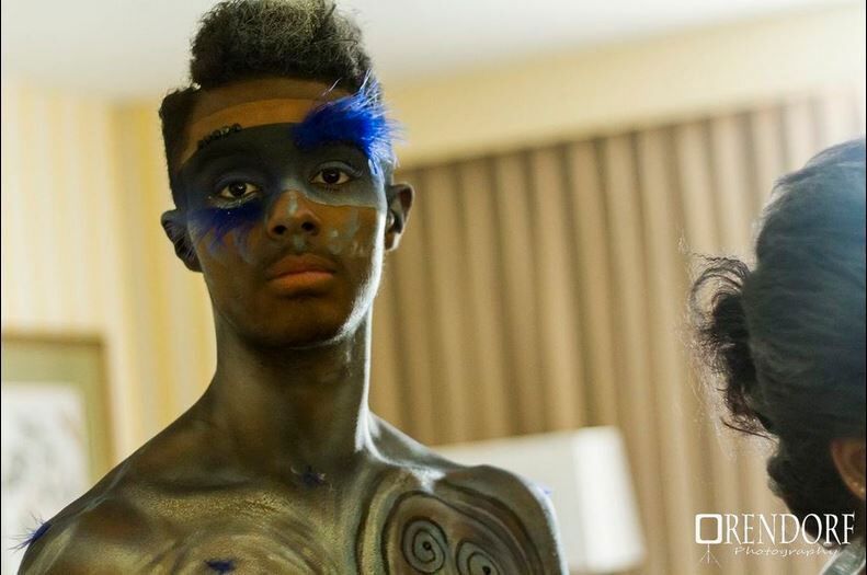 Person with blue and black body paint poses, indoors, looking at the viewer.