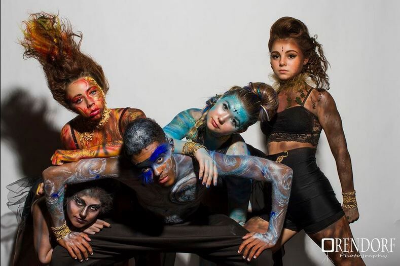 Five people posed with body paint, on a light background. Arms intertwined, with various colors and designs.