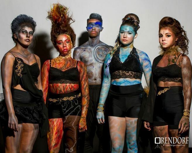 Five people with body paint and themed hairstyles pose; one is shirtless, all in front of a white backdrop.