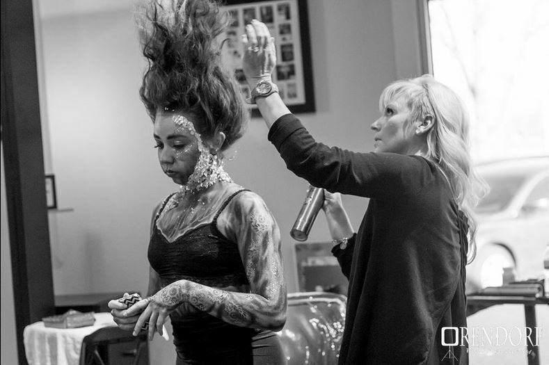 Woman in elaborate makeup being styled by a person with blonde hair holding a spray can. Indoors, looking at a mirror.