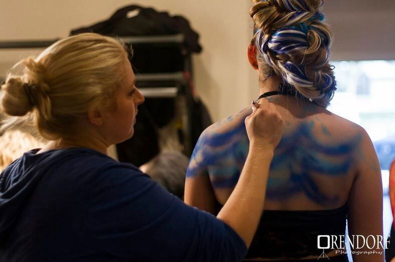 Woman applies blue body paint to another woman's back. Indoors, focused work.