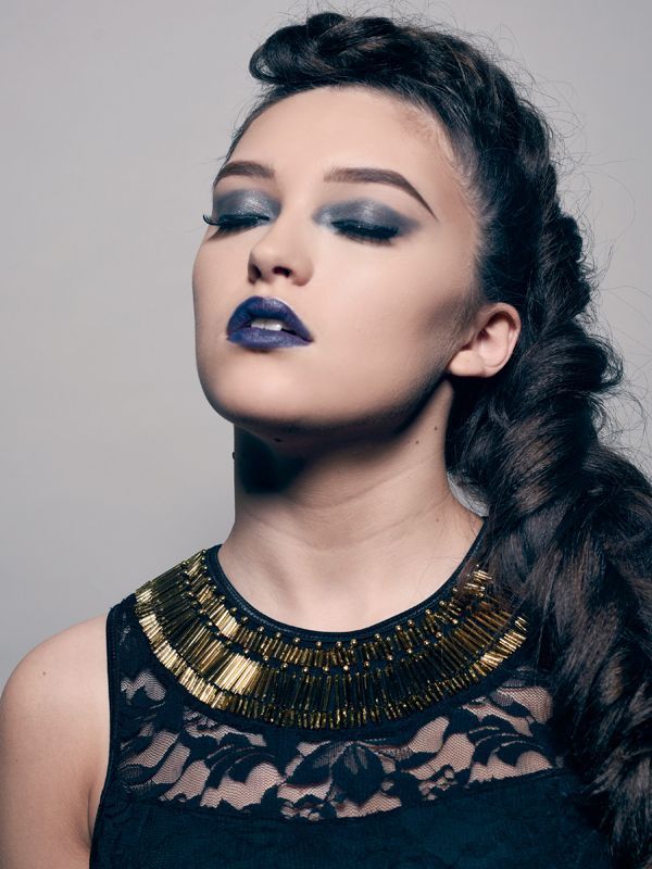 Woman with dark hair, eyes closed, wearing dramatic blue makeup and a gold-accented necklace.