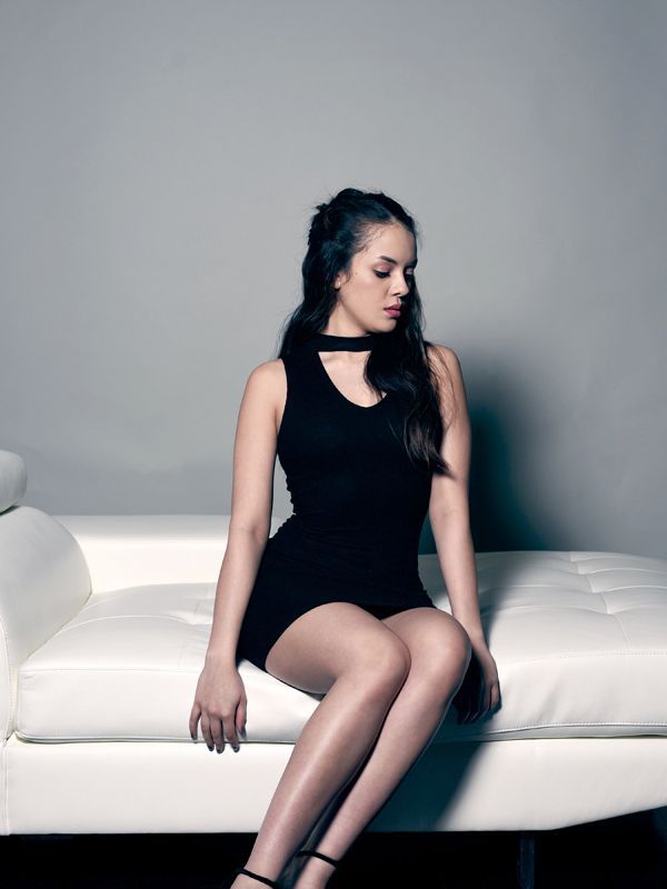 Woman in black dress sits on white couch, looking down. Gray background.