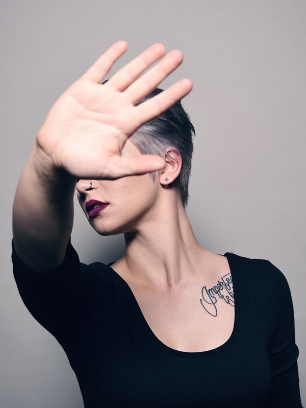 Woman with short hair, covering her face with an open hand. A tattoo is visible on her chest.