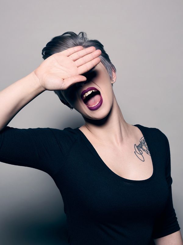 Woman with short, gray hair shielding her eyes, mouth open wide, dark lipstick, tattoo, black top.