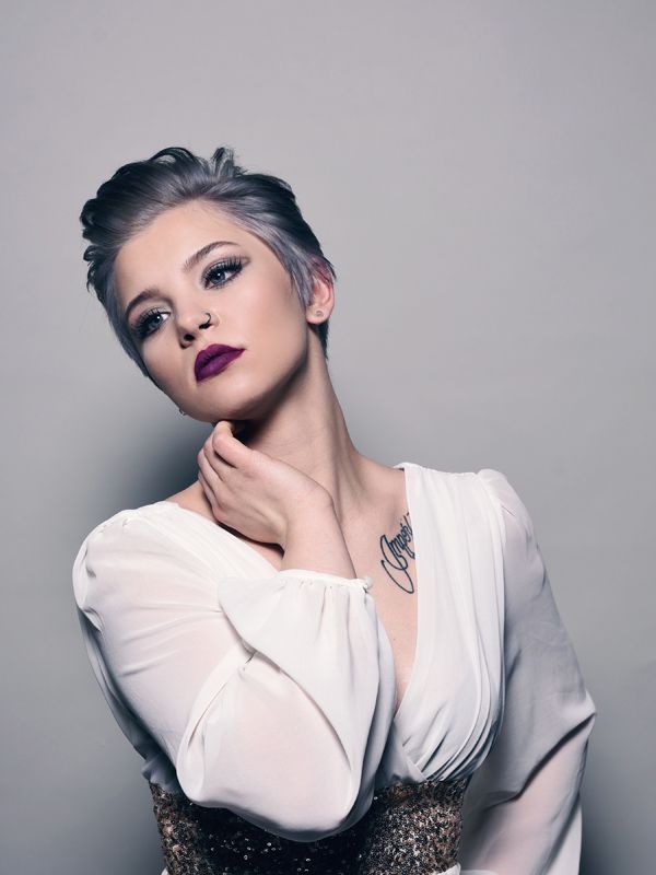 Woman with short, silver hair and dark lipstick, wearing a white dress and touching her neck, a tattoo on her chest.