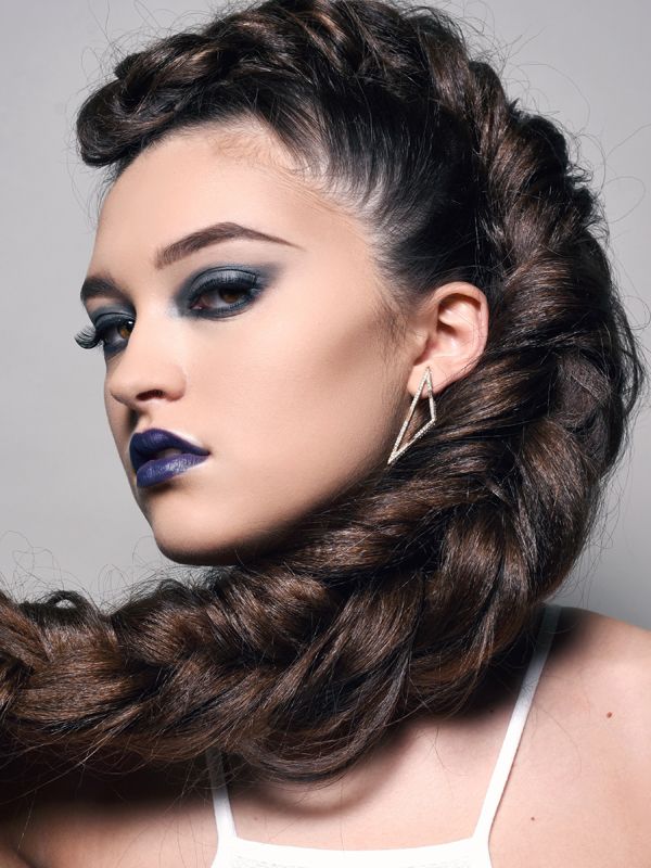 Woman with dark hair in a braided updo, wearing dark eye makeup and lipstick, a white tank top, and a diamond earring.