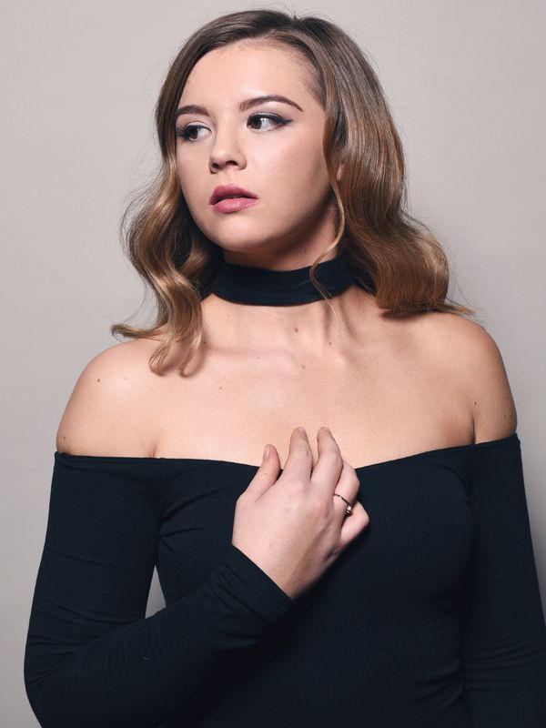 Woman wearing black off-the-shoulder top and choker, looking to the side, hand on chest.