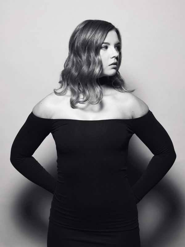 Woman in a black off-the-shoulder top, arms at sides. Soft waves, neutral background.