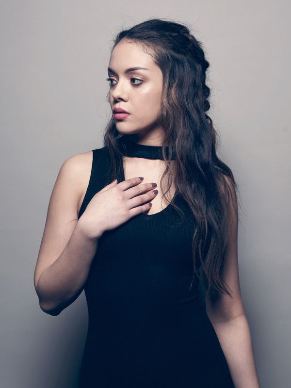 Woman in black dress, holding hand on chest, looking to the side with dark braided hair.