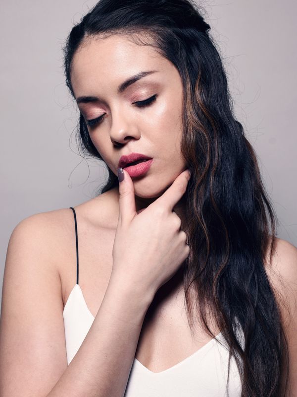 Woman with long wavy hair, pink lipstick, touching her lip, wearing a white spaghetti strap top, eyes closed.