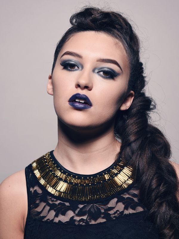 Woman with dark makeup, wearing a black lace top and a gold-beaded collar, hair in a side braid, looking upwards.