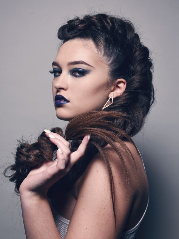 Woman with dark hair styled in an updo and holding a long braid. Wearing dark eyeshadow and purple lipstick.