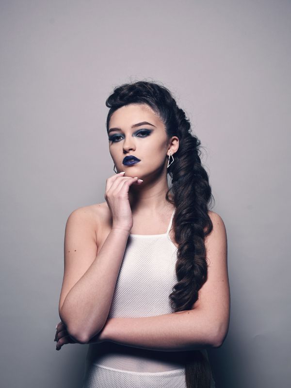 Woman with dark braided hair and makeup poses, wearing a white dress and earrings, against a gray backdrop.