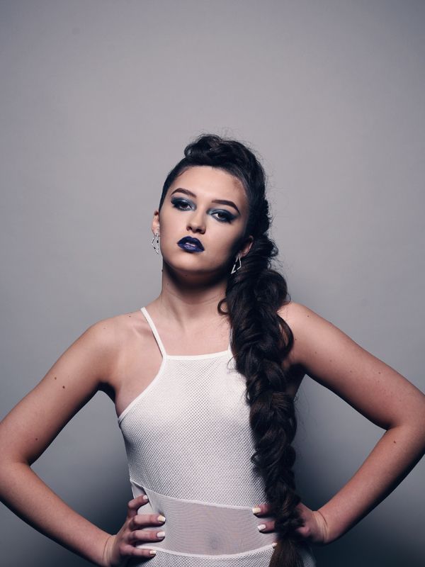 Woman with dark braided hairstyle and dark lipstick, wearing a white dress, hands on hips.