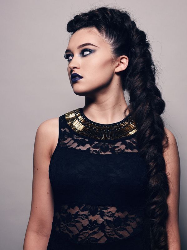 Woman with dark makeup and long braided hair, wearing a black lace top and gold-accented collar, looking to the side.