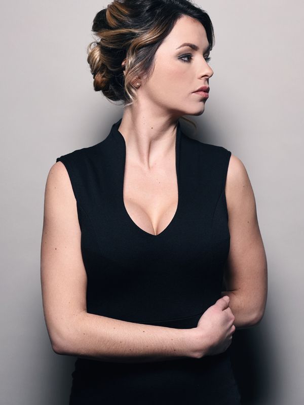 Woman in black sleeveless dress, looking to the side, arms crossed. Updo hairstyle. Gray background.