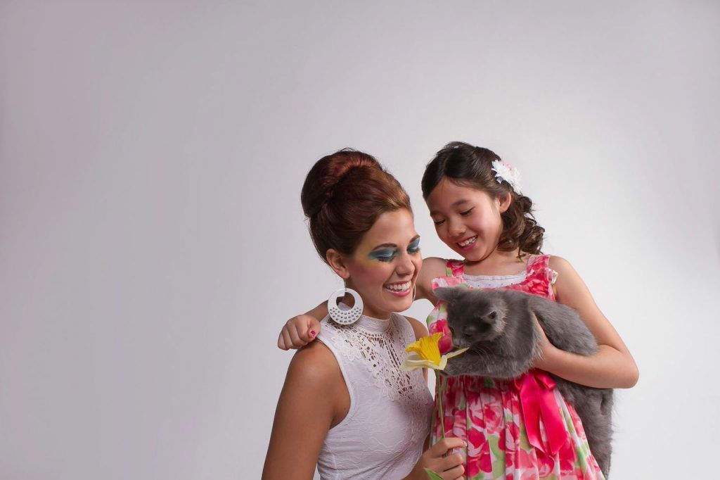 Woman and girl smiling, holding a gray cat with a flower, against a white background.