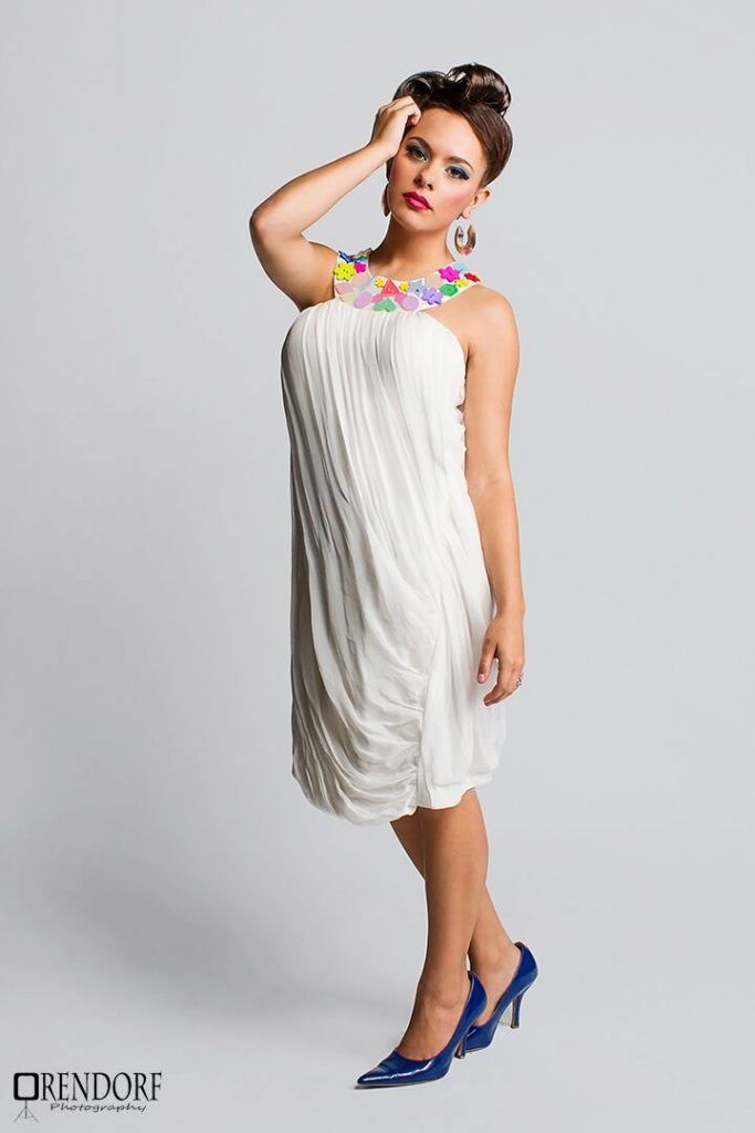 Woman in white dress, colorful necklace, blue heels, hand on head, studio setting.