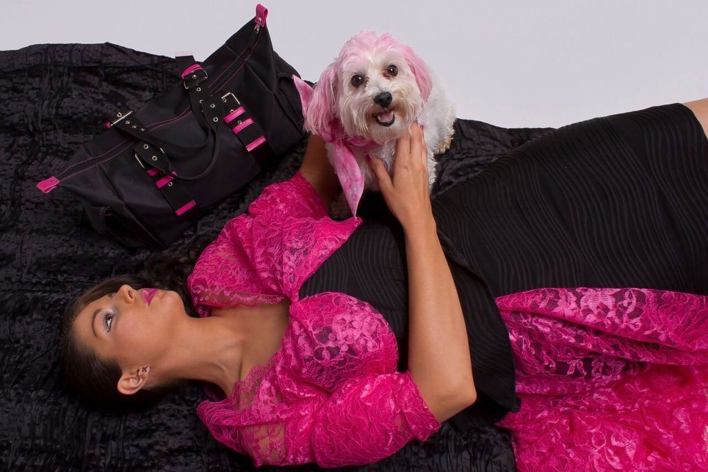 Woman in black dress with pink accents, lying with small dog wearing a pink accessory, on black textured surface.