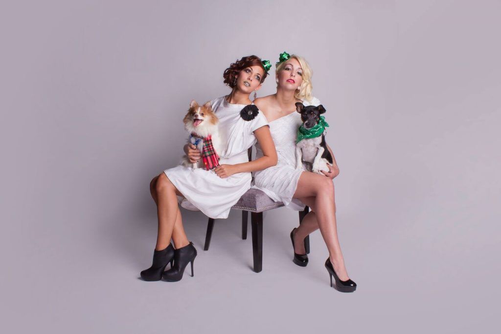 Two women in white dresses sit holding dogs, all with bows, against a grey backdrop.