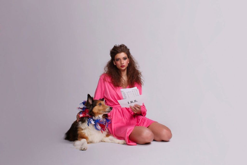 Woman in pink robe sits next to dog with blue and red decoration, holding papers, looking concerned on a white background.