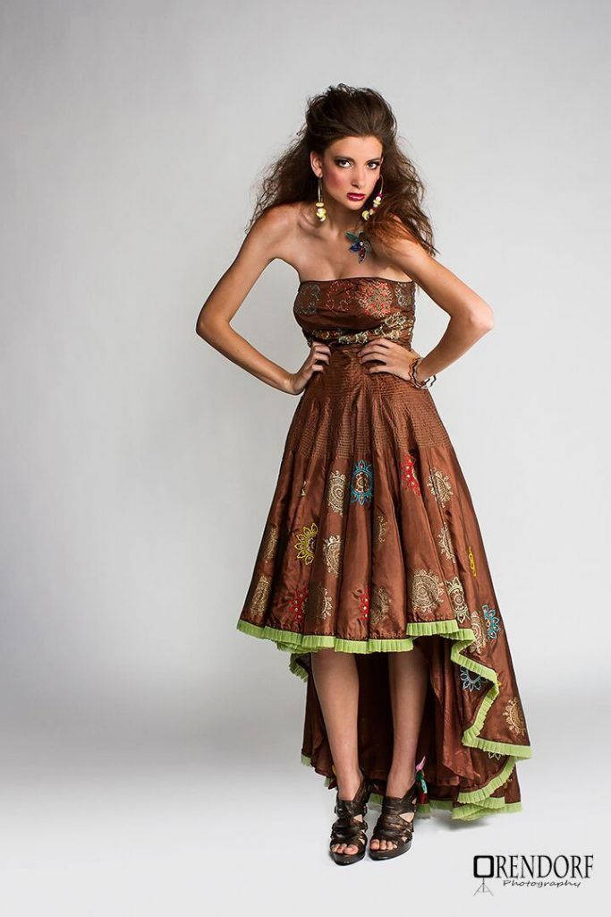 Woman in a brown strapless dress with colorful floral accents, high-low hem, and green trim, posing.